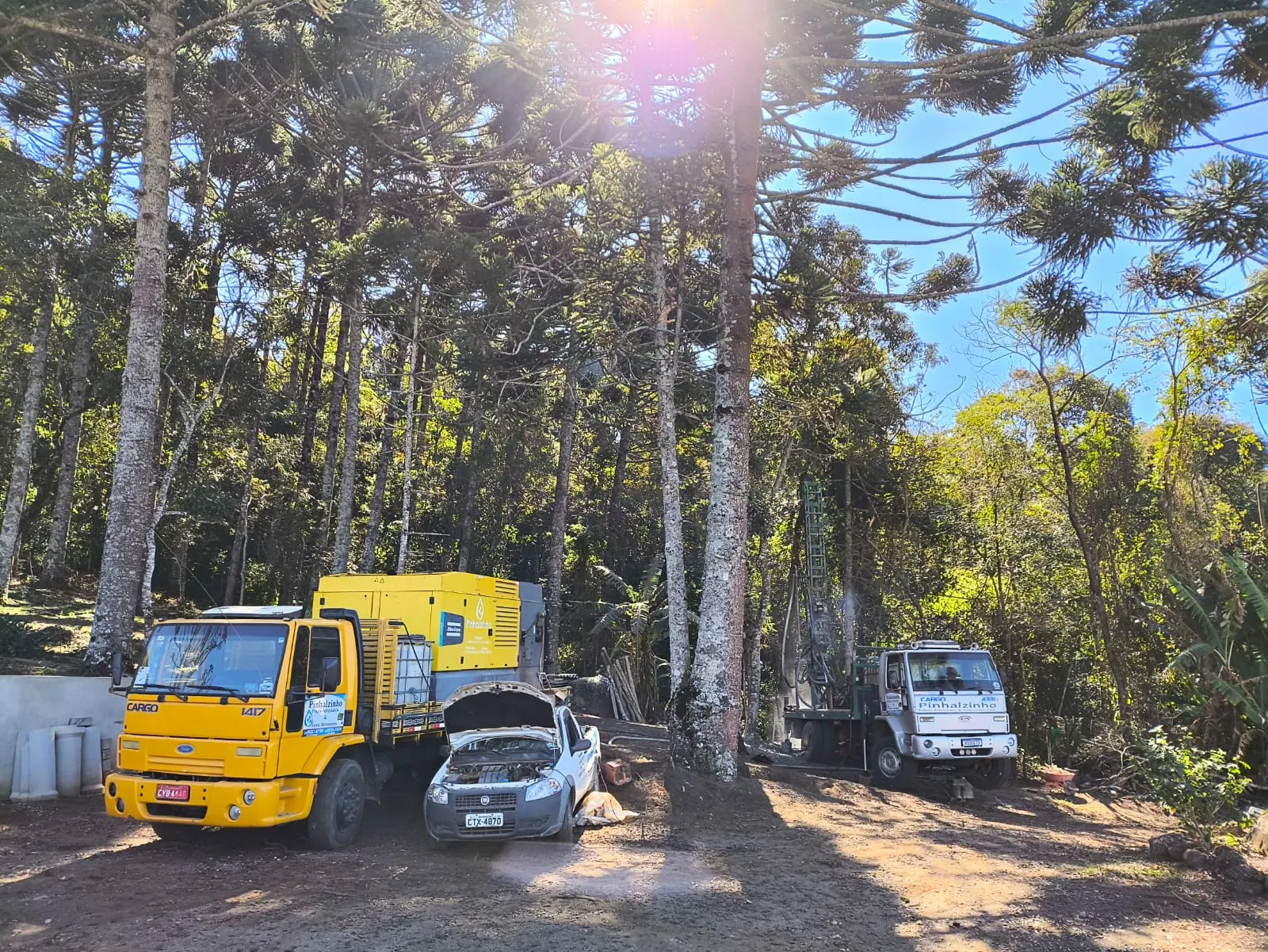 Veículos e equipamentos da Pinhalzinho Poços reunidos em uma área arborizada.